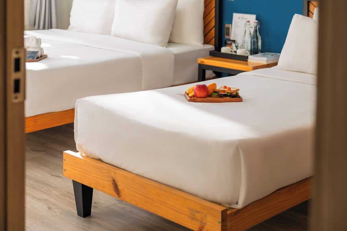 Habitación de hotel con dos camas, pared azul decorada con plumas blancas y faroles, mesita de noche de madera con una lámpara, y una bandeja de frutas sobre la cama.