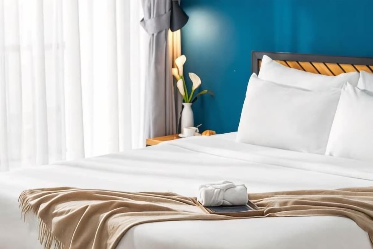 Una cama bien hecha con una colcha blanca y un plaid color beige, un cabecero de madera y una mesita de noche con un jarrón de flores.