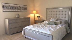 Ein Schlafzimmer mit einem großen Bett, einem Schminktisch, einem Nachttisch mit Lampe und einem an der Wand hängenden abstrakten Gemälde.