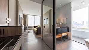Een moderne hotelkamer met een kitchenette, een zithoek met een bank en een eettafel, een badkamer met een grote spiegel en een raam met uitzicht op de stad.