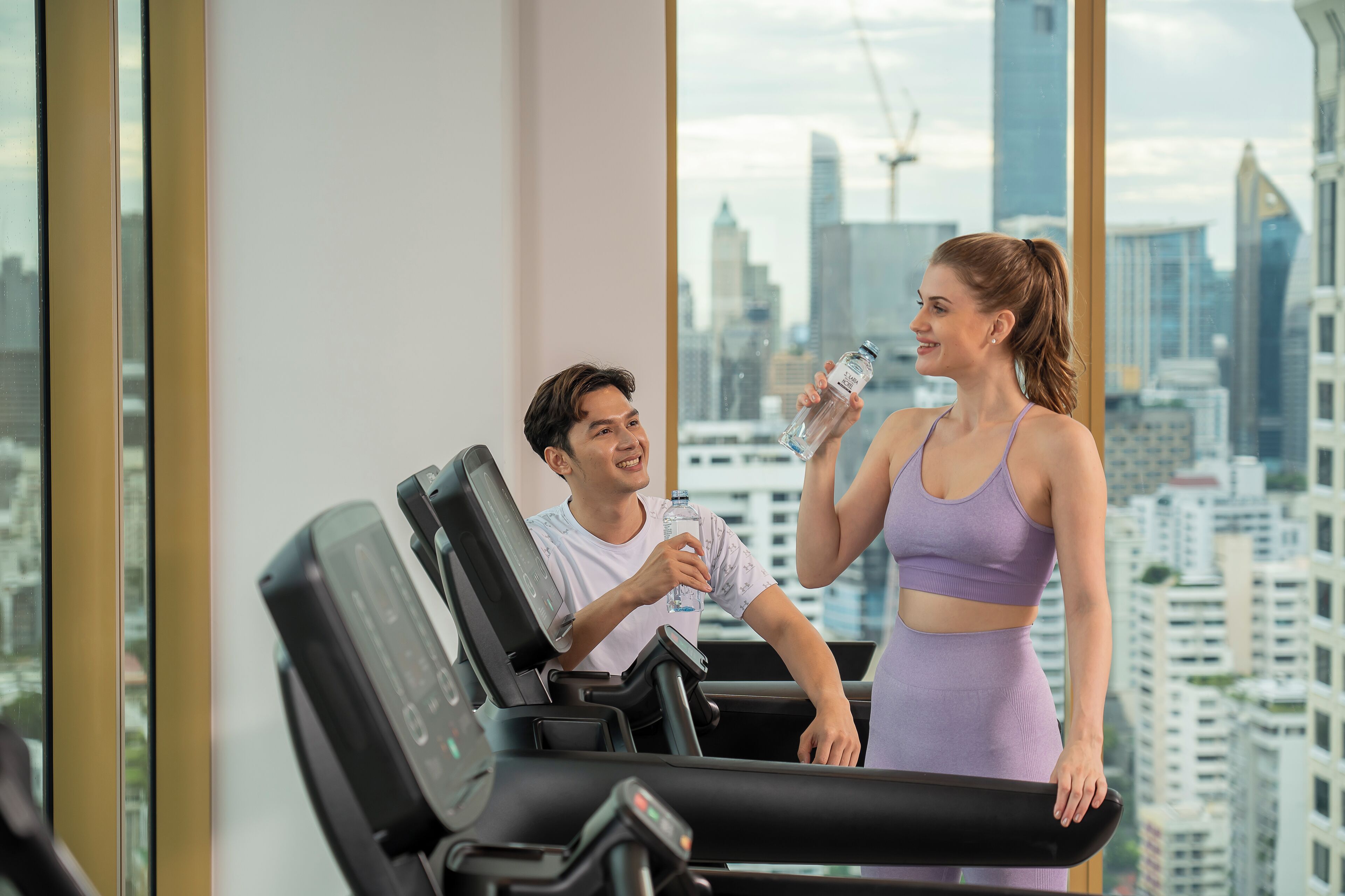 A gym with a city view, a treadmill, and a person holding a water bottle.