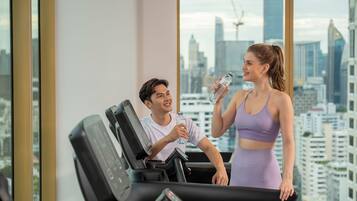 A gym with a city view, a treadmill, and a person holding a water bottle.