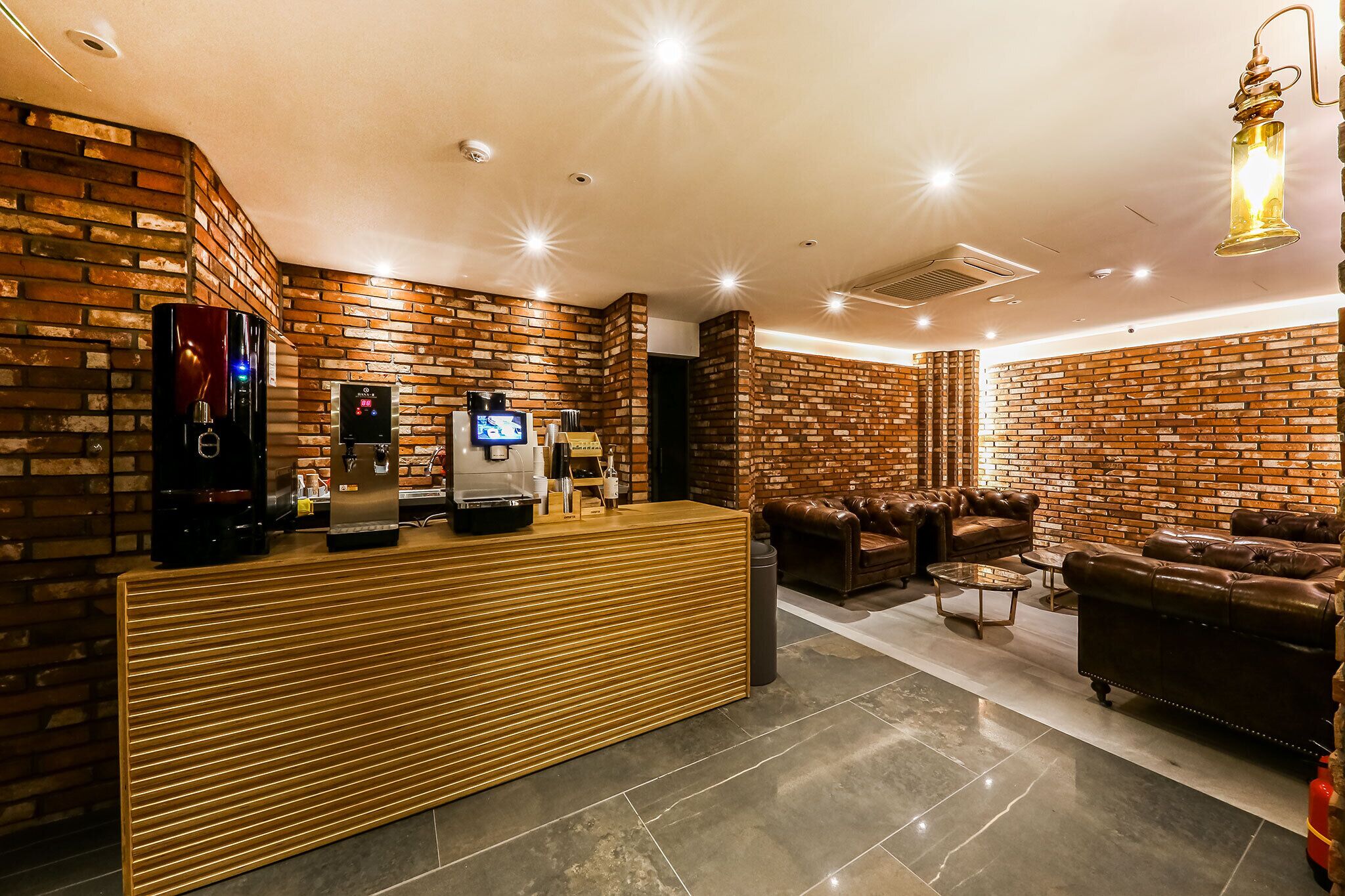 Zona de recepción con mostrador de madera, máquina de café y pantalla digital. Hay sofás de cuero y suelo de baldosas.