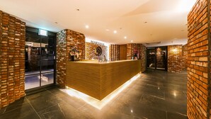 Zona de recepción con un mostrador de madera, paredes de ladrillo y una puerta de cristal que da al exterior.