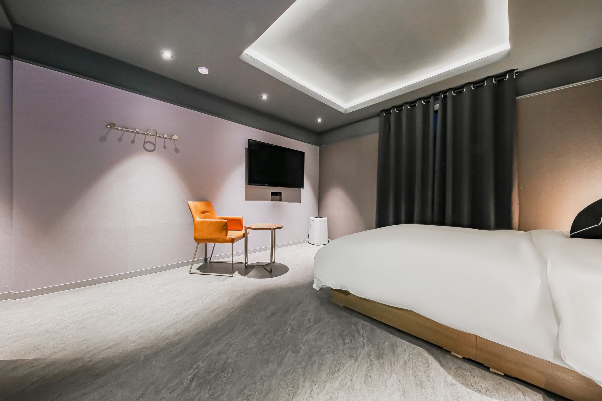 A modern hotel room with a large bed, a flat-screen TV mounted on the wall, a small table and chair, and a ceiling light fixture.