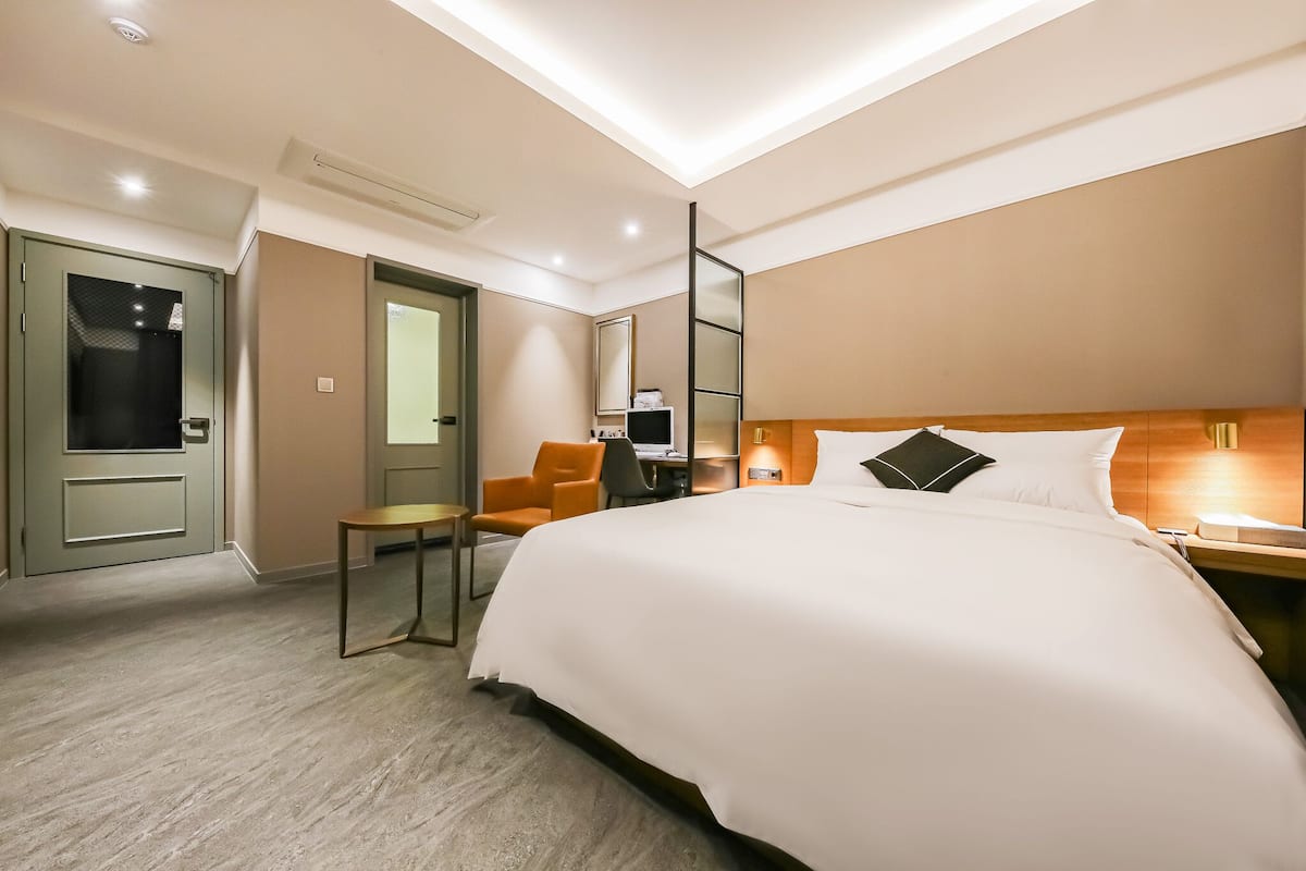 A hotel room with a large bed, a desk with a chair, a mirror, and a door.