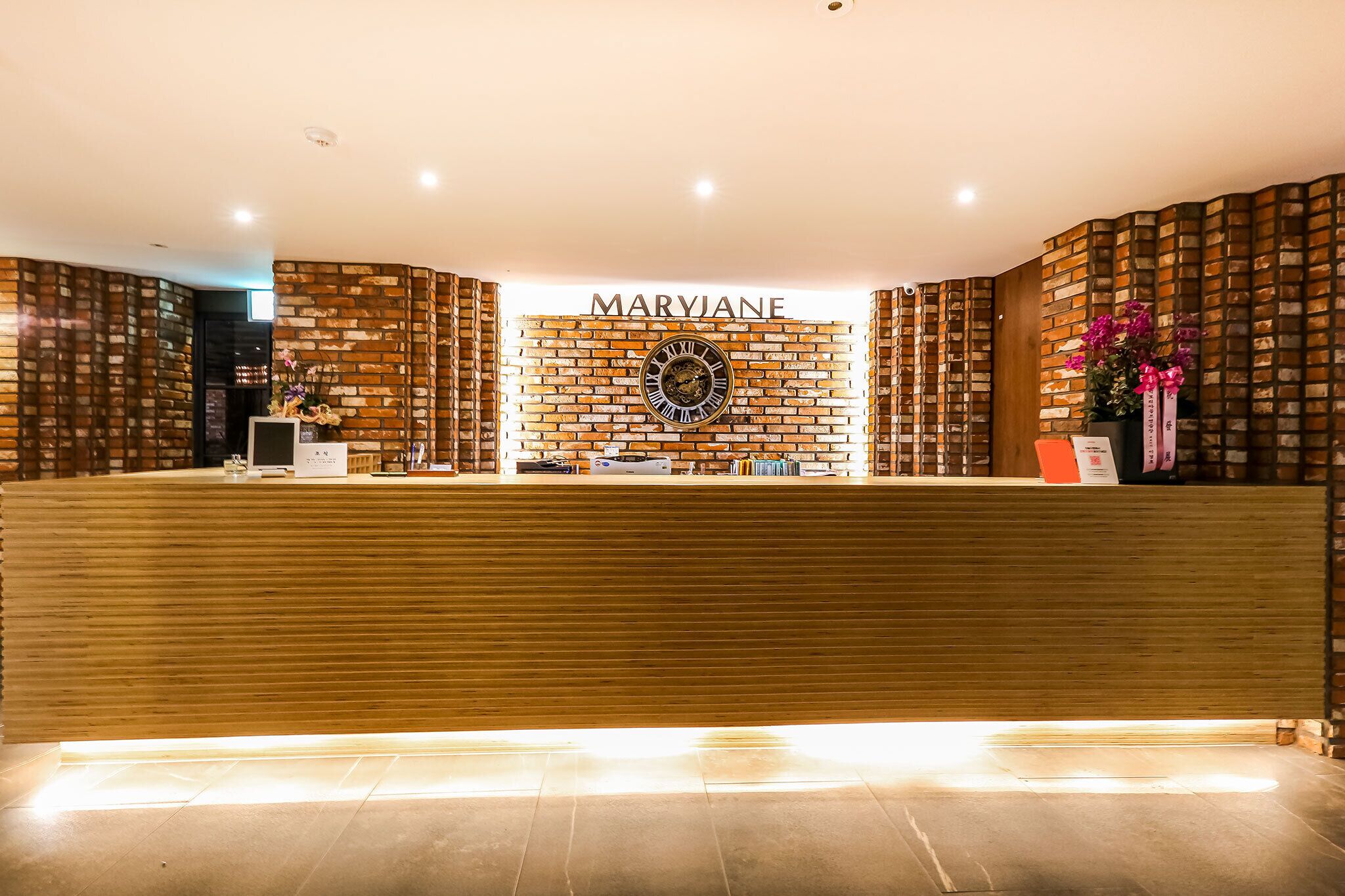 Zona de recepción con un mostrador de madera, una pared de ladrillo con el nombre "MARYJANE" y un reloj.