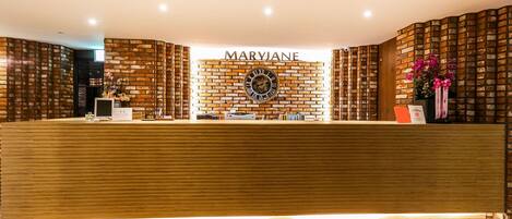 Zona de recepción con un mostrador de madera, una pared de ladrillo con el nombre "MARYJANE" y un reloj.