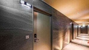 Un pasillo moderno con una puerta de ascensor metálica, un cartel sobre ella y varias puertas a lo largo del corredor.