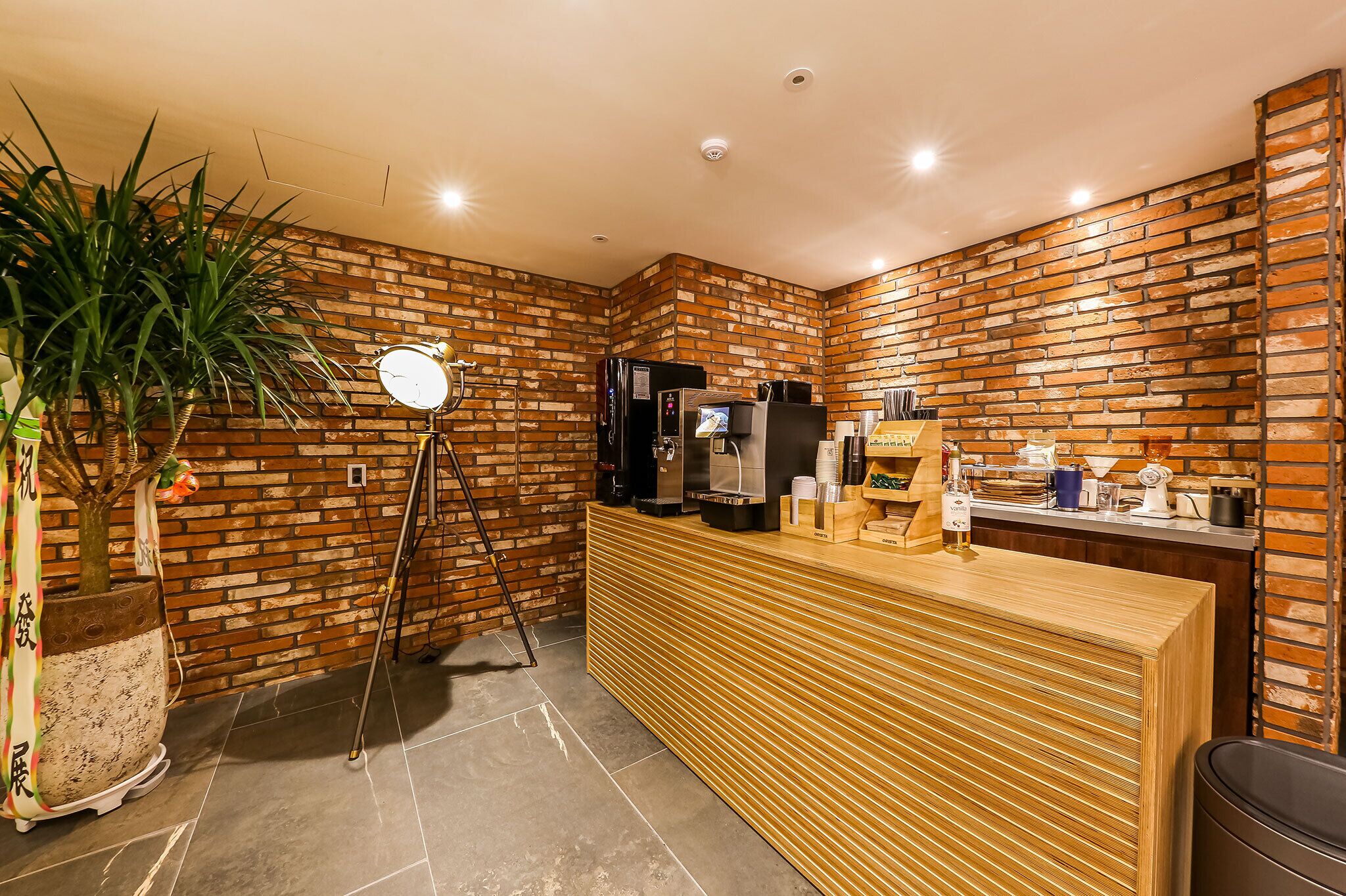 A reception area with a brick wall, a wooden reception desk, a coffee machine, and a potted plant.