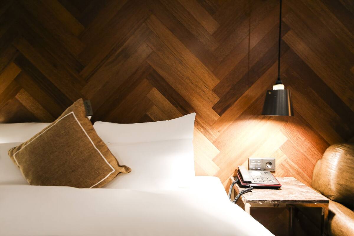 A room with a herringbone wooden ceiling, a bed with white linens and a brown pillow, a bedside table with a lamp, a telephone, and a radio.