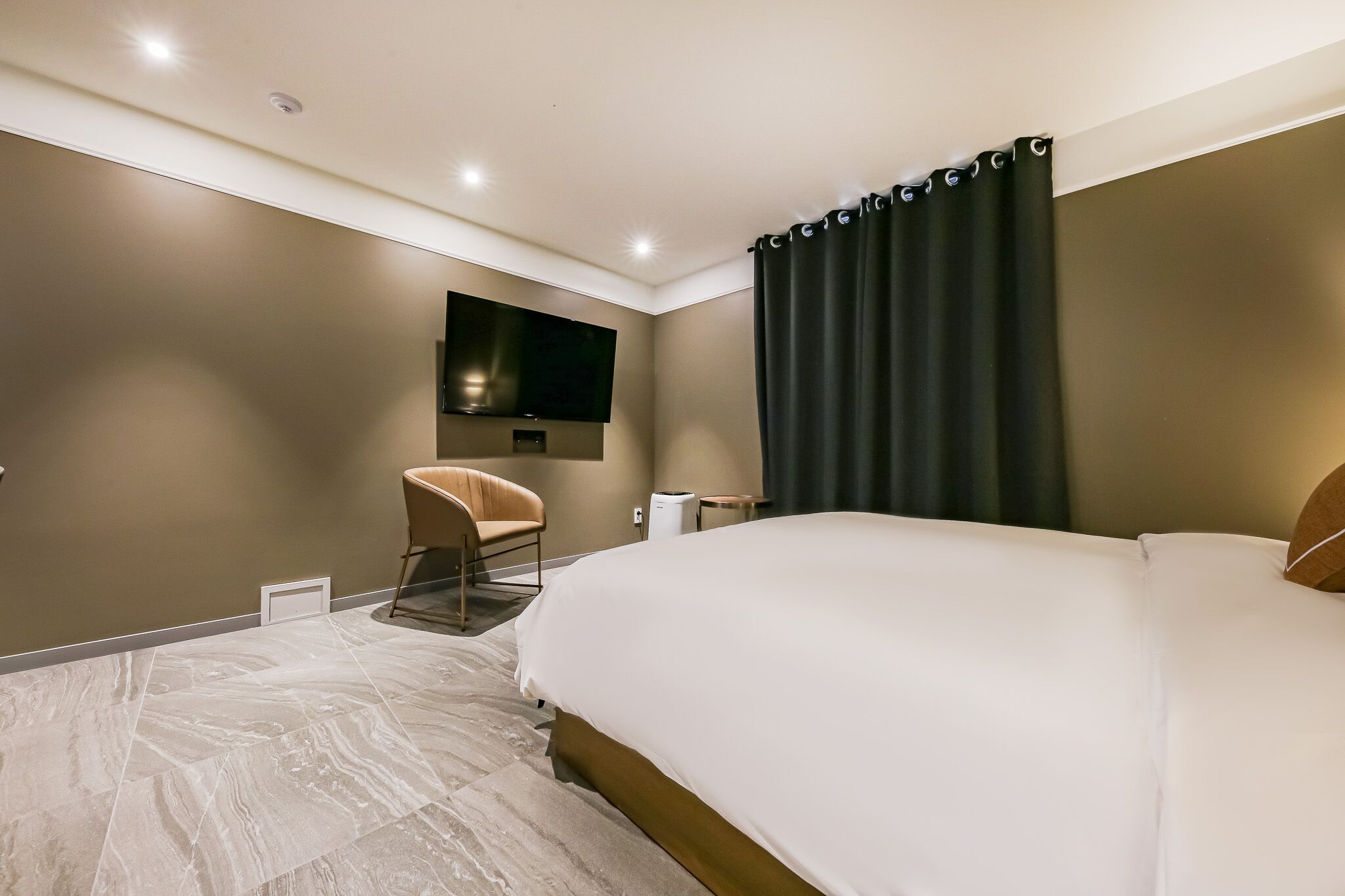 A modern hotel room with a large bed, a flat-screen TV mounted on the wall, a chair, and a small table.