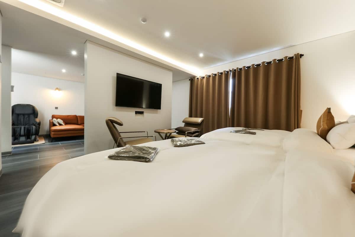 A hotel room with a large bed, a TV mounted on the wall, a seating area with a chair and a small table, and a sofa in the background.