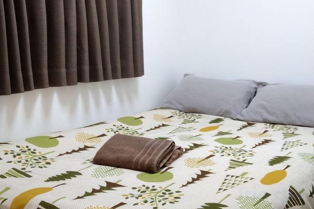A bed with a patterned bedspread, a folded brown towel, and two pillows.