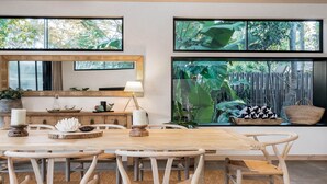 Un comedor con mesa y sillas de madera, un espejo grande y vistas a un jardín tropical a través de amplios ventanales.