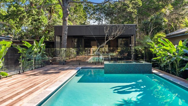 Un área de piscina moderna al aire libre con cubierta de madera, rodeada de plantas tropicales y una estructura de edificio contemporánea.