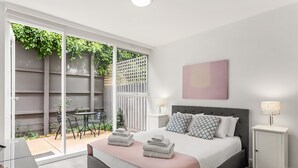 Quarto com cama, mesas de cabeceira, abajur e vista para um pátio externo.