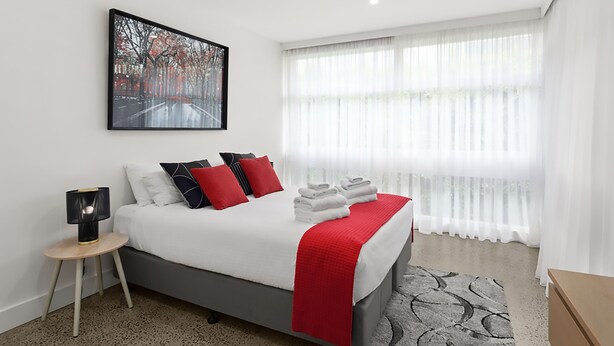 Quarto de hotel moderno com uma cama grande, mesa de cabeceira, luminária e janela com cortinas finas.