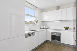 A modern kitchen with white cabinetry, a built-in dishwasher, a stainless steel oven, and a window with a view of the outside. - Brighton By Sea with sea glimpses (Brighton)