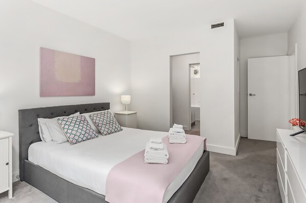 A modern bedroom with a bed, bedside tables, a TV, and a bathroom door.