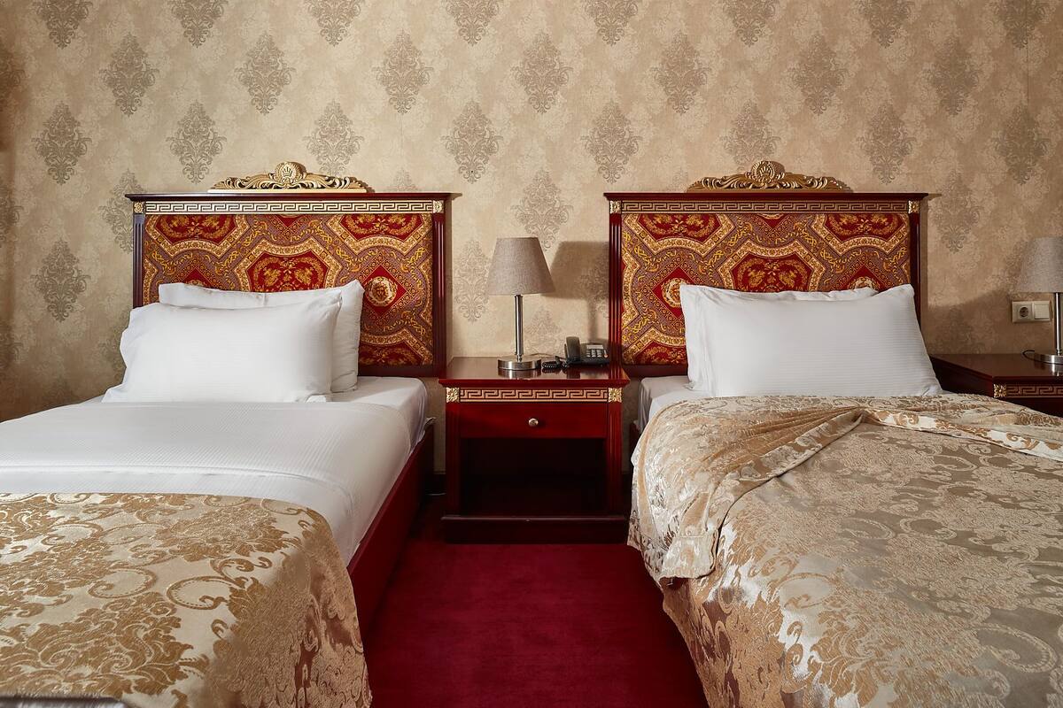 A hotel room with two beds, each with a red headboard and a white pillow. There is a nightstand between the beds with a lamp and a phone. The walls have a patterned wallpaper.