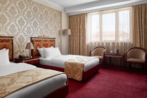 A hotel room with two beds, a desk, two chairs, and a large window with curtains. - Hotel Gold (Skopje)