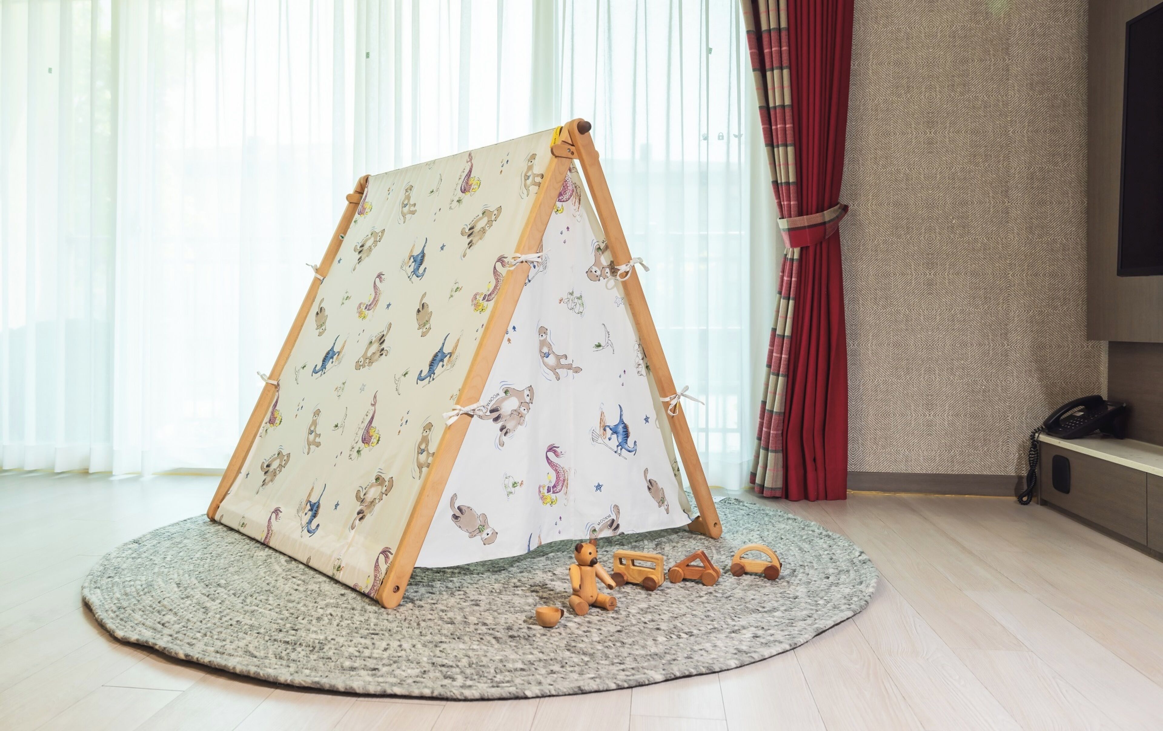Uma área de recreação com uma tenda de brinquedo de madeira estampada com padrões de animais, um tapete redondo e brinquedos de madeira no chão.
