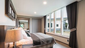 Moderni hotellihuone, jossa on suuri sänky, yöpöytä lampun kanssa, ikkuna verhoilla ja näkymä ulos.