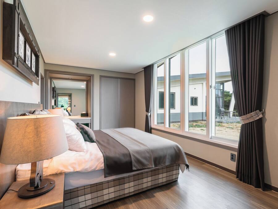 A modern hotel room with a large bed, a bedside table with a lamp, a window with curtains, and a view of the outside.