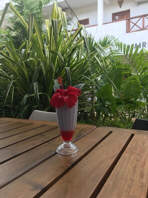 A wooden table with a clear glass drink in a tall glass, surrounded by lush green foliage. - 27 Cafe Zanzibar Airport Hotel (Zanzibar Town)
