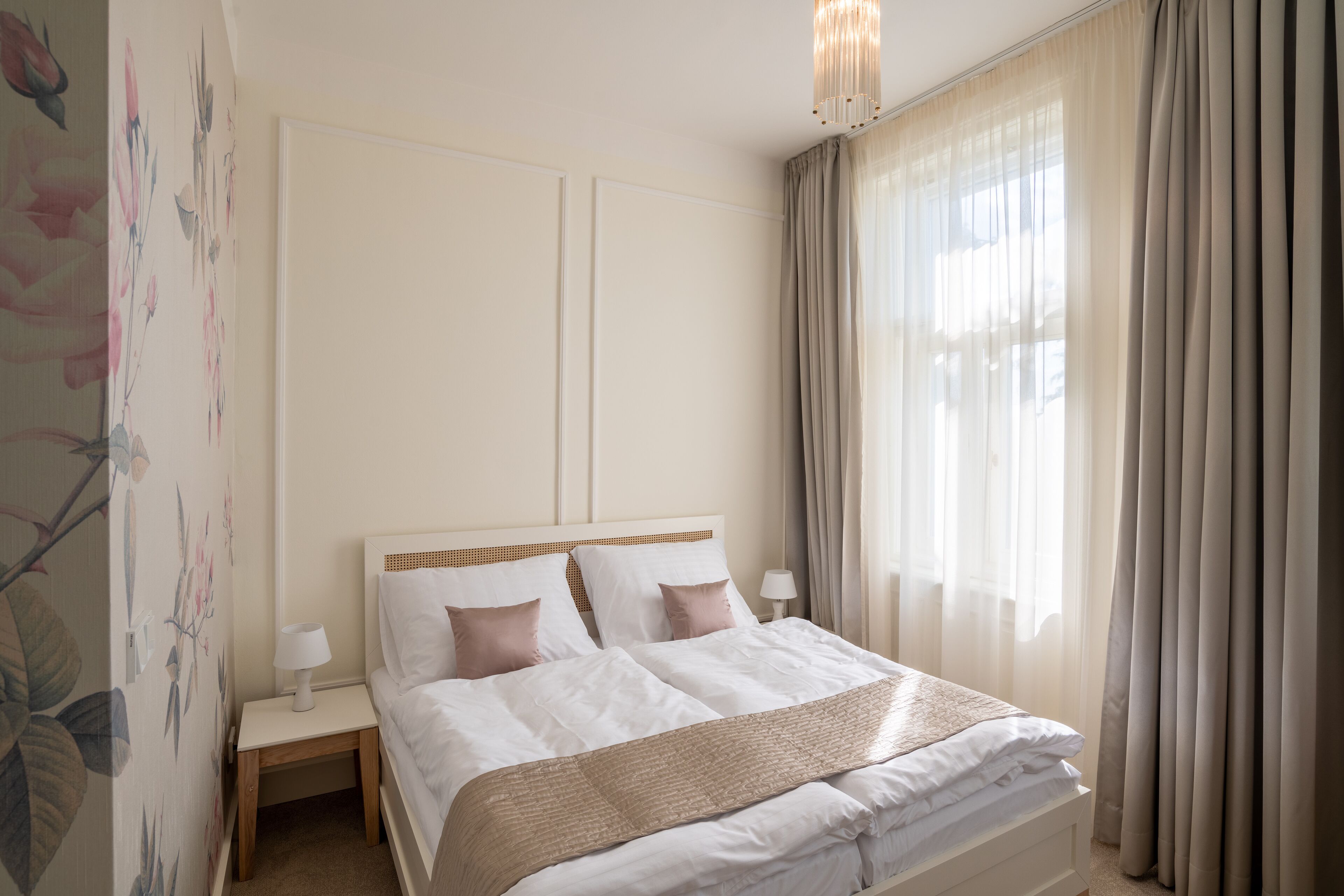 Una cama bien hecha con sábanas blancas y una manta de color beige, una mesita de noche con una lámpara y un estampado floral en la pared.