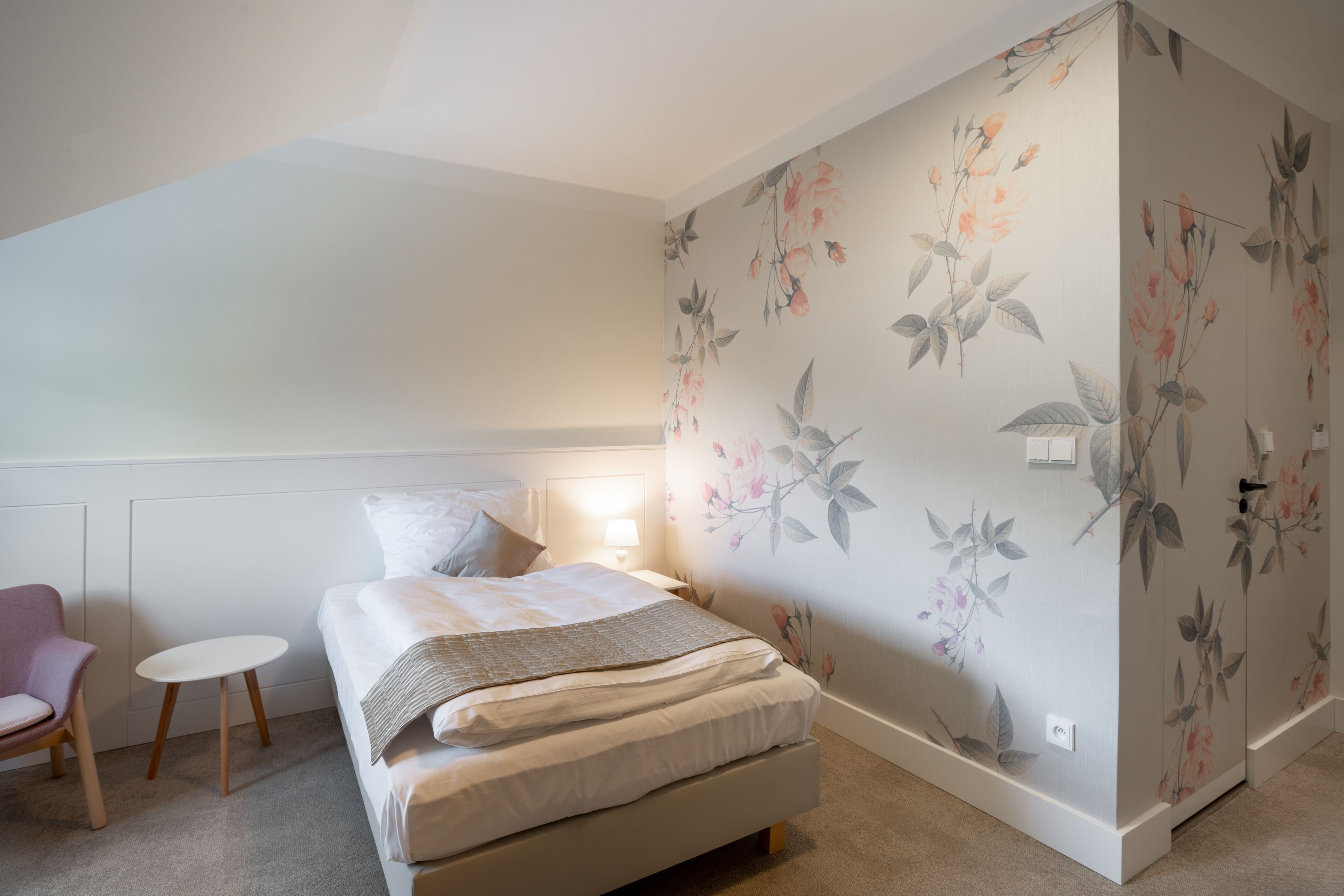 Un dormitorio con una cama, una silla, una mesita y papel tapiz con motivos florales.
