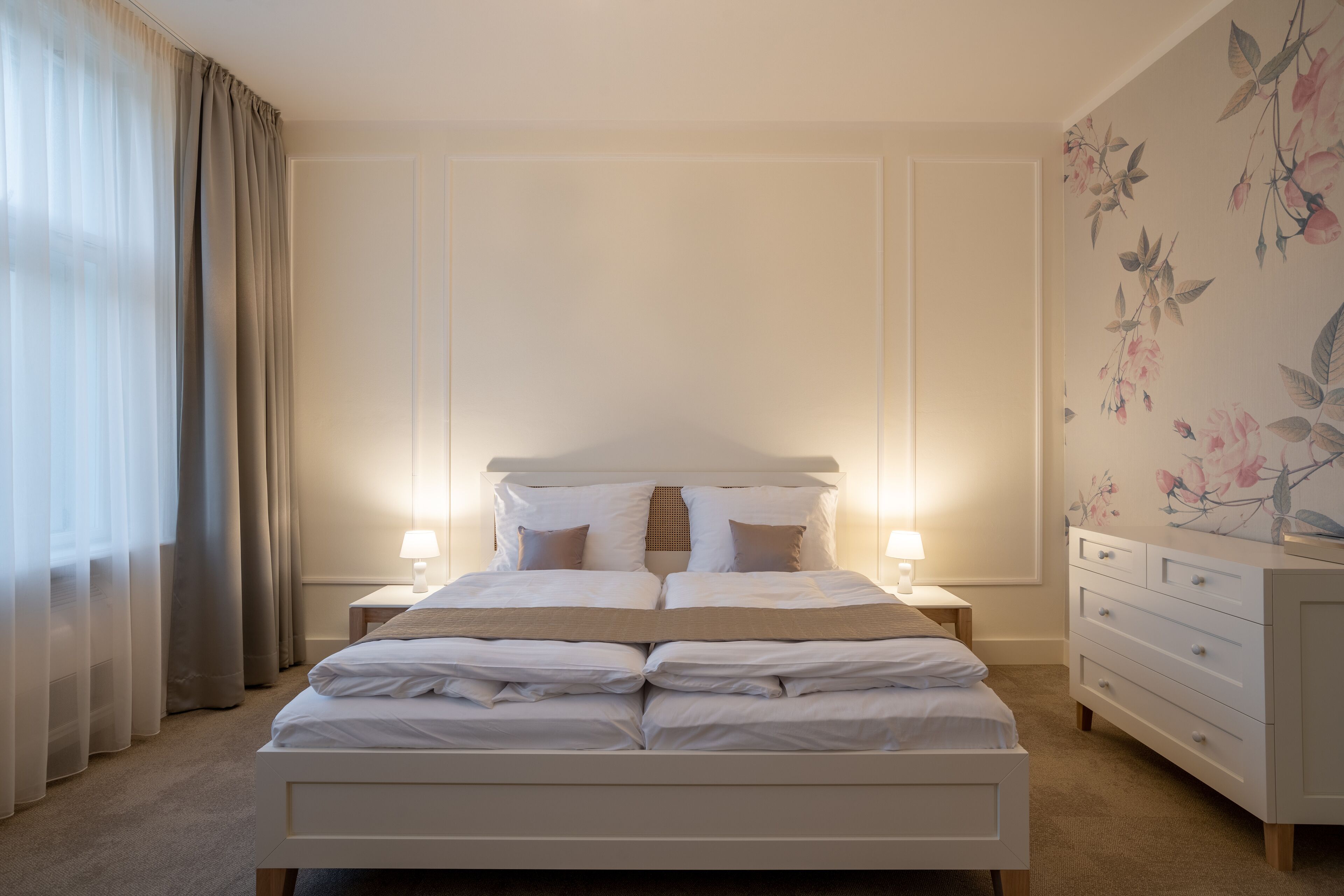 A neatly made bed with white linens and two bedside lamps, a dresser with drawers, and a floral wallpaper on the wall.