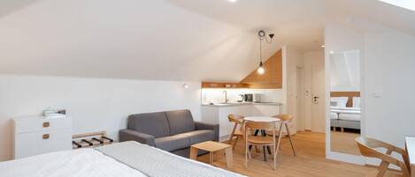 Una habitación moderna y bien iluminada con una cama, un sofá gris, una pequeña mesa de comedor y una kitchenette al fondo.