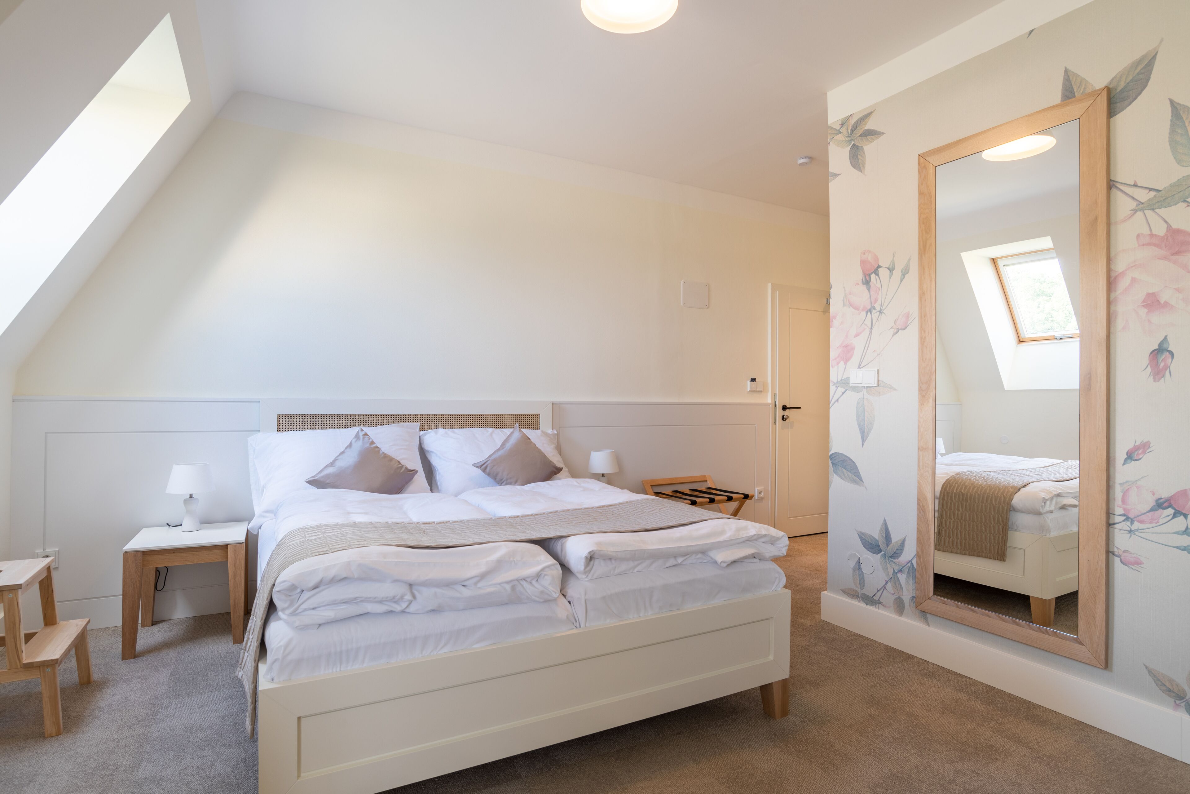 A bedroom with a large bed, a nightstand, a wooden chair, a mirror, and a window with a view of the sky.