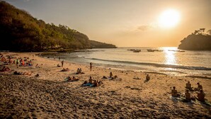 Une plage où des gens se prélassent au soleil et se baignent, un ciel dégagé et le soleil qui se couche sur l’eau.