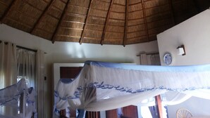 A room with a thatched roof, a bed with a white and blue canopy, a wooden chair, and a wall-mounted clock. - Parkside Safari Lodge (Pakwach)