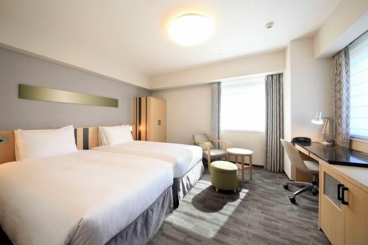 Ein Hotelzimmer mit zwei Betten, einem Schreibtisch, einem Sessel, einem kleinen Hocker und einem Fenster mit Vorhängen.