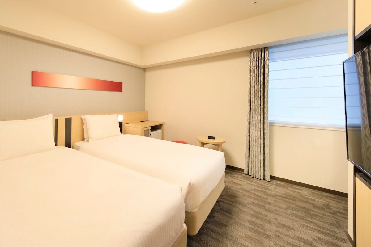 A hotel room with two beds, a desk, a TV, and a window with curtains.