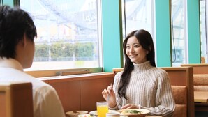 Una persona sentada a una mesa con comida, en una habitación con ventanas amplias y vistas a un puente.