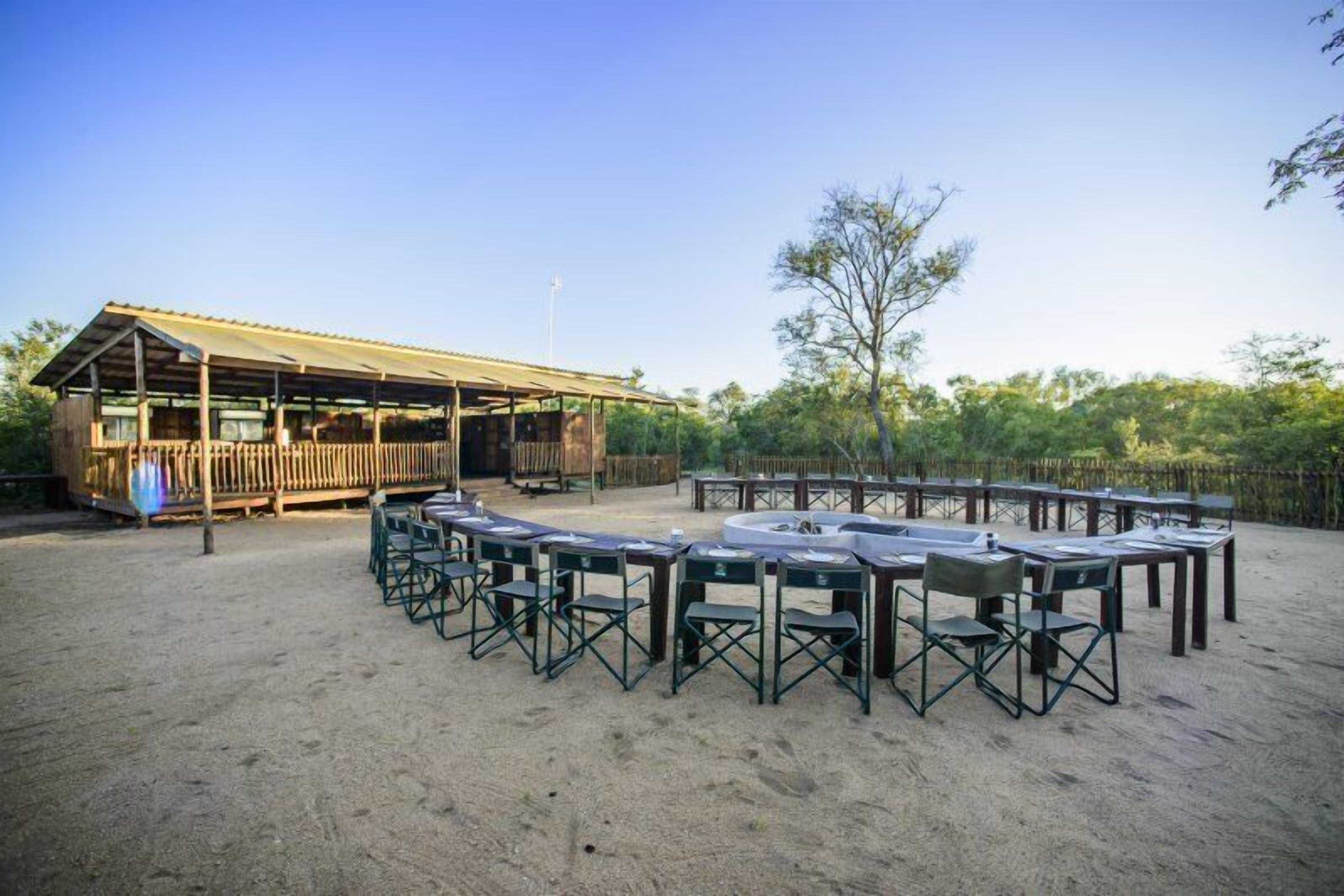 An outdoor seating area with tables and chairs arranged in a semi-circle, a wooden building with a covered roof, and a sandy ground.