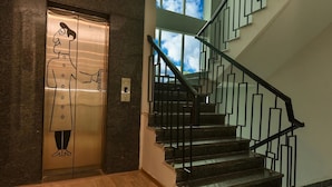 An elevator with a cartoon character, a staircase with black railings, and a view of the sky through the windows. - Mavens White Artemis Hospital Road Gurgaon (Gurugram)
