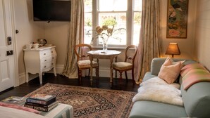 Et værelse med en sofa, et lille bord med stole, et fjernsyn ophængt på væggen, et vindue med gardiner og et billede på væggen.
