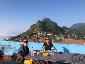 A dining table with food and drinks, set against a backdrop of a mountainous landscape. - The Hotel Bandipur (Bandipur)