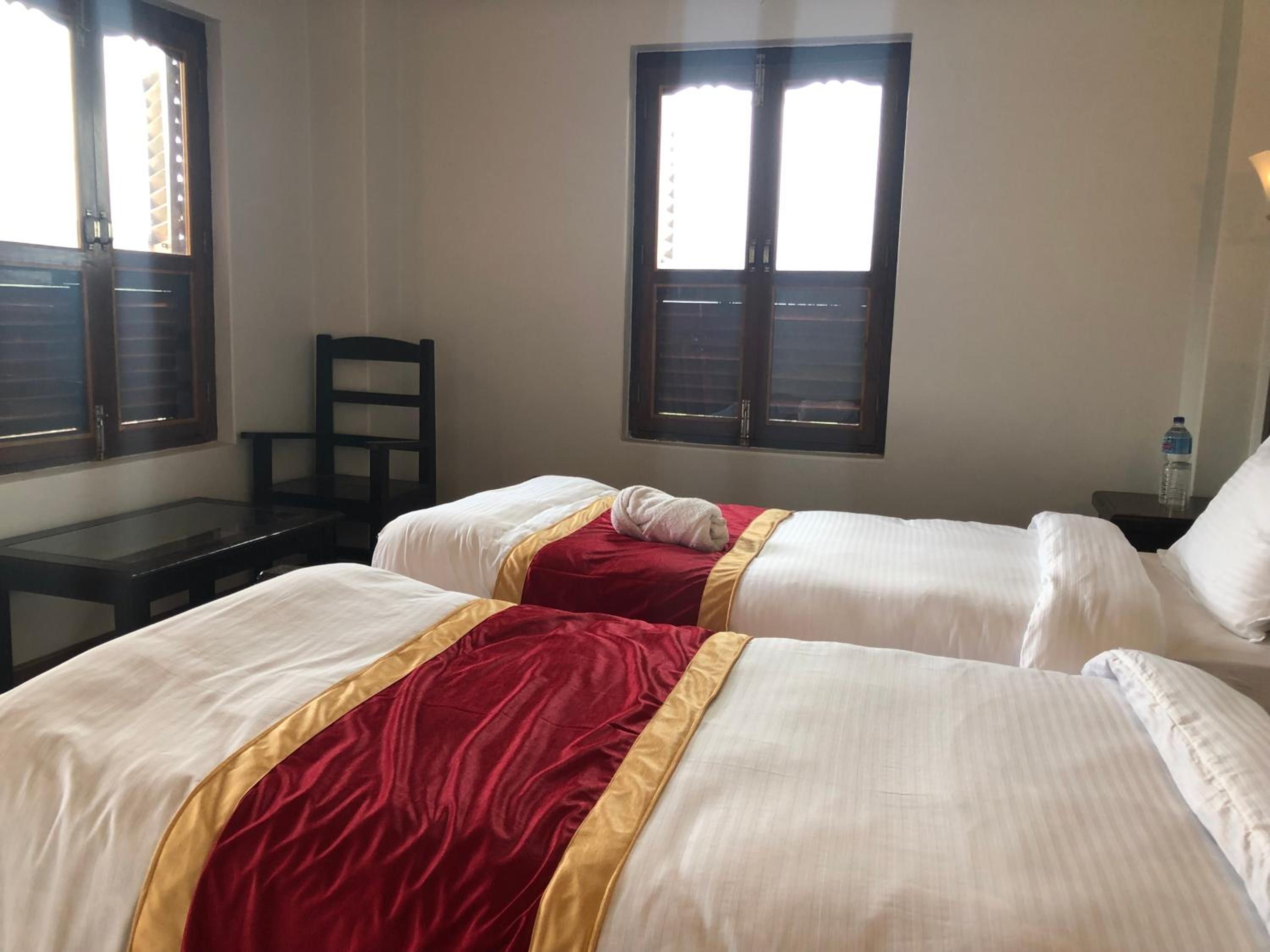 A hotel room with two beds, a desk, and a chair. There are windows with blinds, a water bottle on the nightstand, and a lamp.