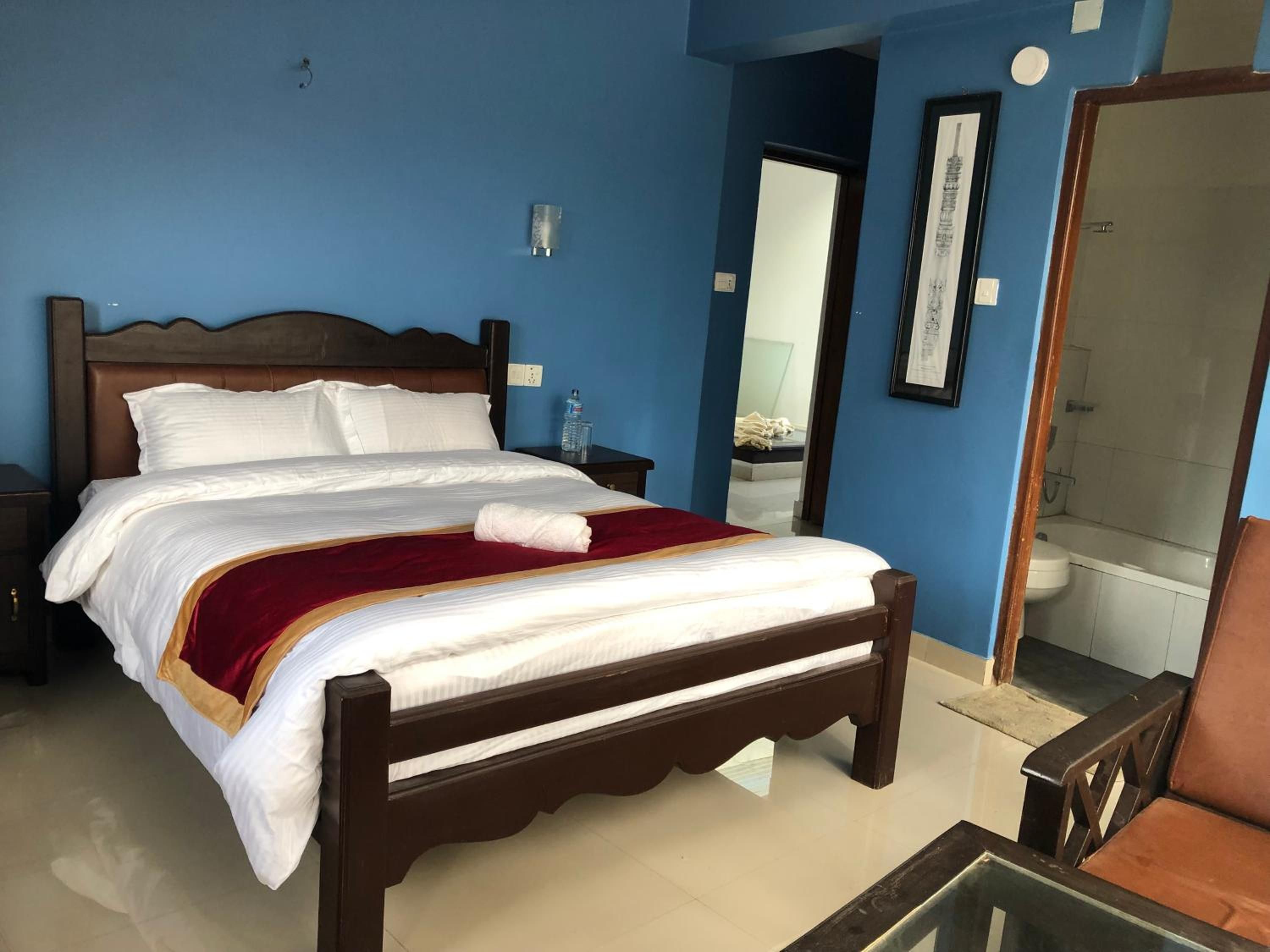 Une chambre d’hôtel avec un lit, une table de chevet, un miroir et une salle de bain visible à travers une porte ouverte.