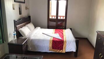 A hotel room with a bed, a bedside table with a water bottle, a glass, and a lamp. There is a window with blinds and a door with a wooden frame.