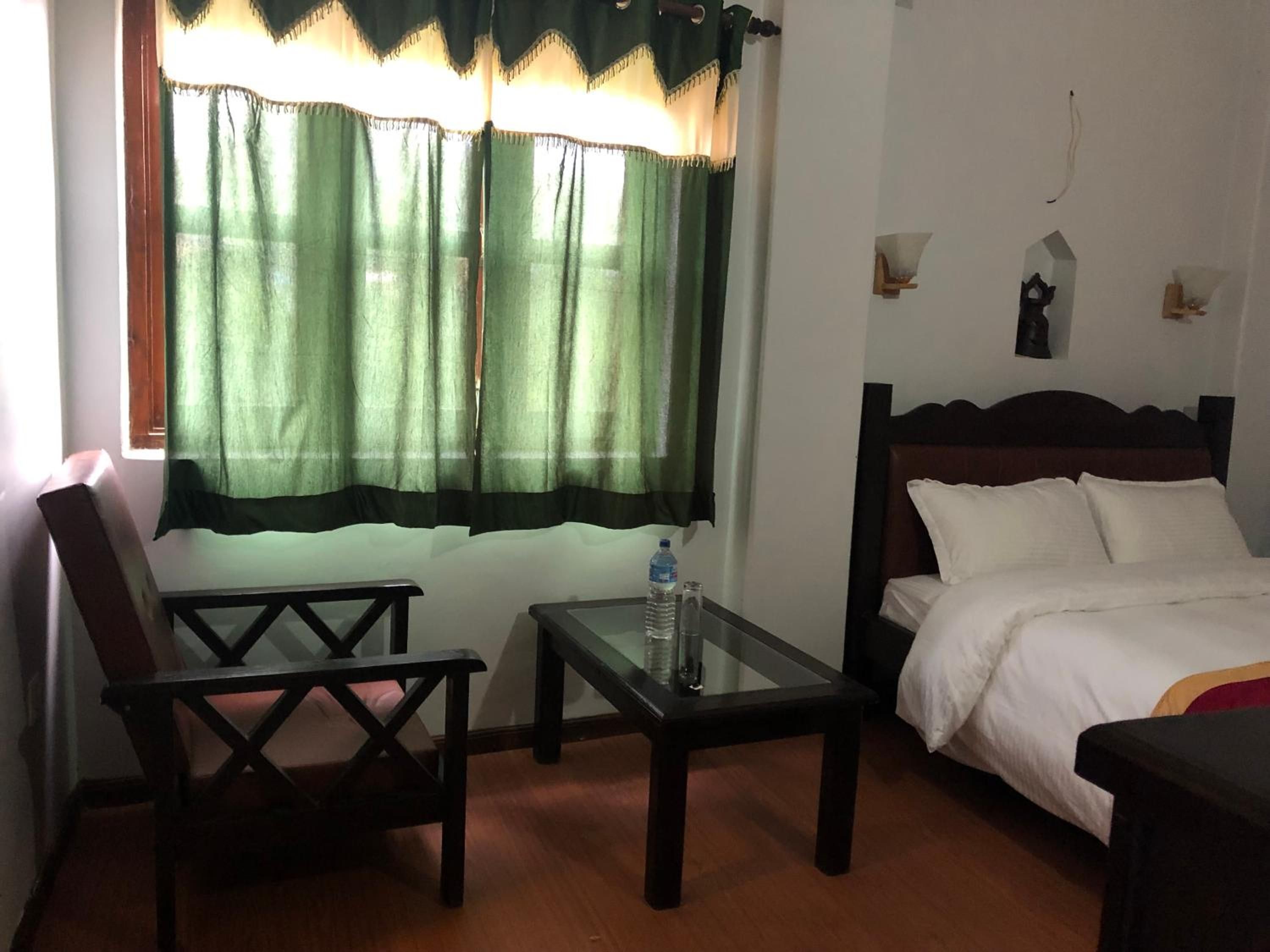 Habitación de hotel con cama, una mesita con una botella, una silla y una ventana con cortinas verdes.