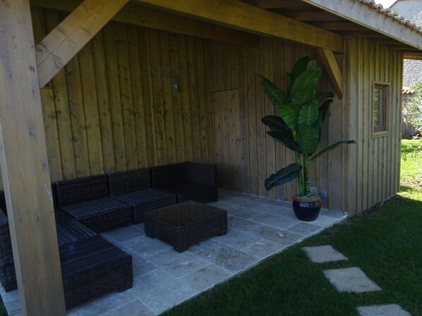 A covered outdoor seating area with a wooden wall, a large potted plant, and a stone-tiled floor. - Domaine la Bastide (Cleyrac)