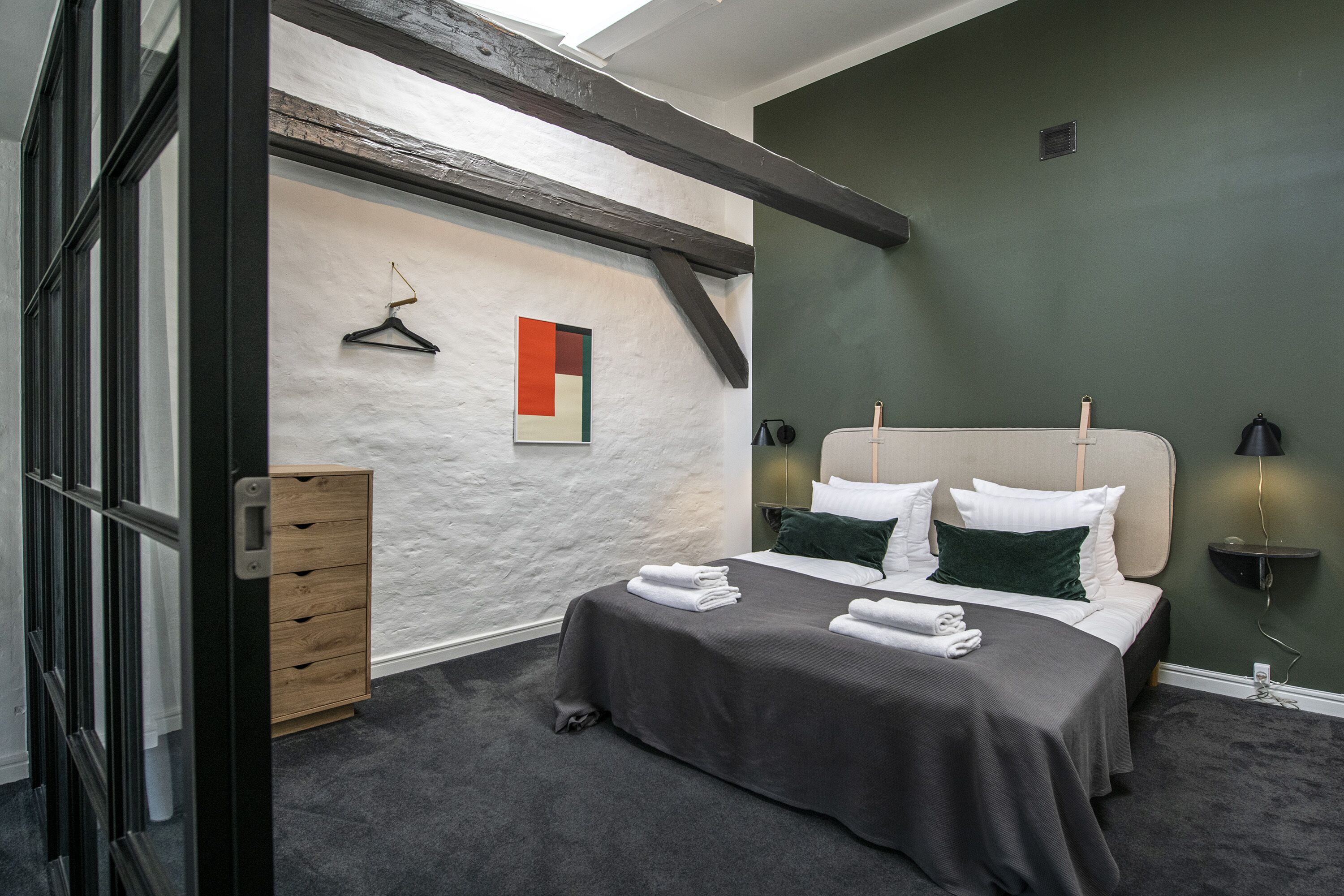 Un dormitorio con cama, cómoda de madera, un cuadro en la pared y una lámpara colgante.